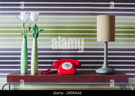 Blumen in grünen Vasen und Retro-Telefon neben Lampe in Reihe auf Holztisch gegen Retro-Tapete platziert Stockfoto