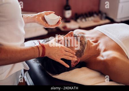 Männer mittleren Alters mit entspannendem Gesicht Massage mit Öl von gesichtslosen Frau in weißer Uniform im Salon Stockfoto