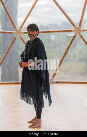 Lächelnder, stilvoller, alter bärtiger Mann in einer Brille, gekleidet in einem trendigen grauen warmen Poncho, der barfuß in einer designten Holzklasse mit Panoramafenster steht Stockfoto
