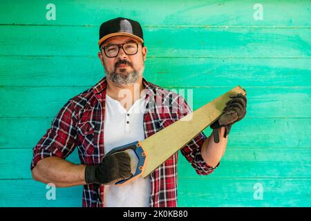 Reifer Rüde in karierten Hemden und Mütze mit Handschuhen, die die Handsäge im Koffer halten, während er vor grünem Holzhintergrund auf die Kamera blickt Stockfoto
