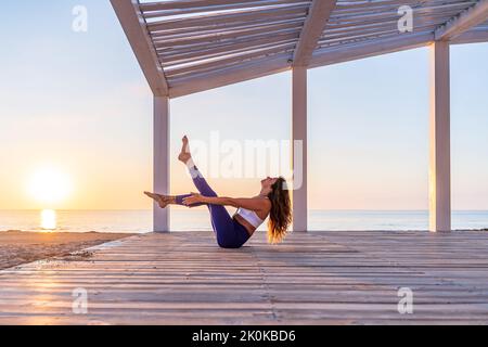 Seitenansicht einer schlanken Frau im aktiven Ohr, die in Navasana Yoga praktiziert Pose auf einer hölzernen Promenade vor dem Hintergrund eines malerischen Sonnenaufgangs am Meer Stockfoto