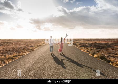 Rückansicht eines unerkennbaren, aufgeregt jungen Paares in stilvoller Kleidung, die Hände hält und auf einer asphaltierten Straße zwischen trockenen vulkanischen Feldern gegen Wolken läuft Stockfoto