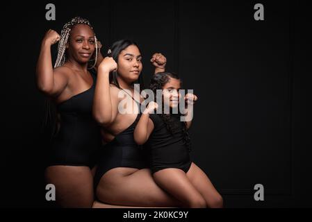 Eine Gruppe sportlicher, multiethnischer, generationsübergreifender Frauen, die vor schwarzem Hintergrund Muskeln zeigen und die Kamera mit einem selbstbewussten Lächeln betrachten Stockfoto