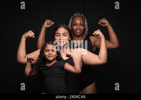 Eine Gruppe sportlicher, multiethnischer, generationsübergreifender Frauen, die vor schwarzem Hintergrund Muskeln zeigen und die Kamera mit einem selbstbewussten Lächeln betrachten Stockfoto