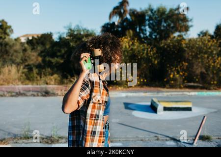 Seitenansicht einer nicht erkennbaren afroamerikanischen Hipsterin mit Afro-Frisur, die auf der Rückseite ein Penny Board auf einer leeren Straße hält und ein sieht Stockfoto