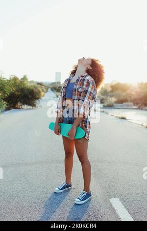 Afroamerikanische Hipsterin mit Afro-Frisur, die mit einem Penny Board auf einer leeren Straße steht und die Augen während des Sonnenuntergangs geschlossen haben Stockfoto