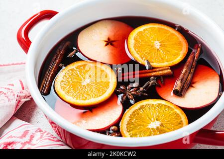 Glühwein mit Orange, Apfel und Gewürzen in einem roten Topf, grauer Hintergrund. Gemütliches Getränk im Herbst oder Winter. Stockfoto