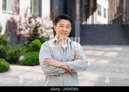 Porträt eines selbstbewussten asiatischen Unternehmers, der mit gekreuzten Armen posiert und die Kamera in der Nähe des Bürogebäudes anschaut Stockfoto