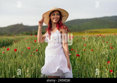 Fröhliches junges Weibchen in Sundress mit roten Haaren, die mit Strohhut wegschauen, der am Sommertag auf dem Feld auf dem Land steht Stockfoto