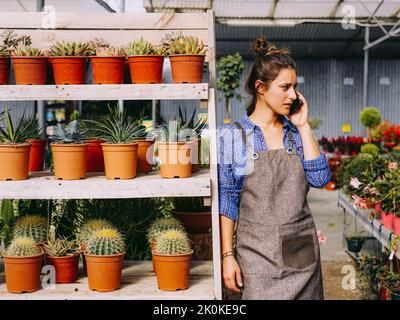 Glückliche Dame im Vorfeld lächelte und beantwortete den Anruf, während sie sich während der Arbeit im Treibhaus mit Sukkulenten auf den Regalen lehnte Stockfoto