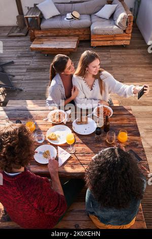 Junge, positive Freunde aus dem Hochwinkel genießen in legerer Kleidung ein köstliches Mittagessen auf der Terrasse und machen Selfie auf dem Smartphone am sonnigen Sommertag Stockfoto