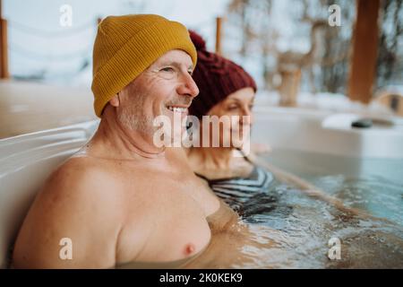 Das ältere Paar mit kitschiger Kappe genießt an kalten Wintertagen gemeinsam die Badewanne im Freien auf der Terrasse. Stockfoto