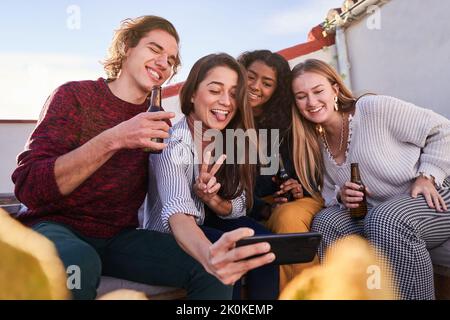 Eine Gruppe von begeisterten jungen multirassischen Freunden in legerer Kleidung, die auf dem Smartphone fratzen und Selfie machen, während sie sich mit Bierflaschen auf der sonnigen Terrasse treffen Stockfoto