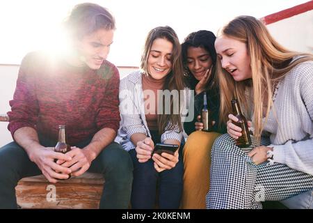 Eine Gruppe von Inhalten junge, vielfältige Freunde, die Bier genießen und auf dem Smartphone surfen, während sie sich am sonnigen Tag auf der Dachterrasse treffen Stockfoto