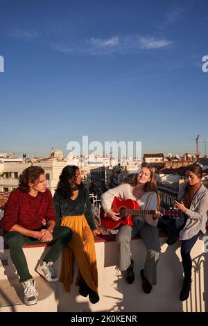 Junge Frau spielt akustische Gitarre zu fröhlichen jungen multirassischen Freunden, während sie auf dem sonnigen Dach feiern Stockfoto