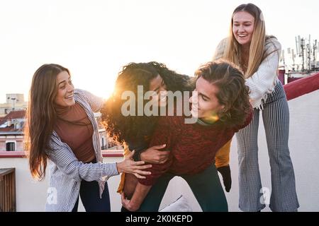 Lächelnd junge männliche Huckepack glücklich weiblich, während Spaß mit Freunden auf dem Dach am frühen Abend Stockfoto