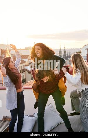 Lächelnd junge männliche Huckepack glücklich weiblich, während Spaß mit Freunden auf dem Dach am frühen Abend Stockfoto