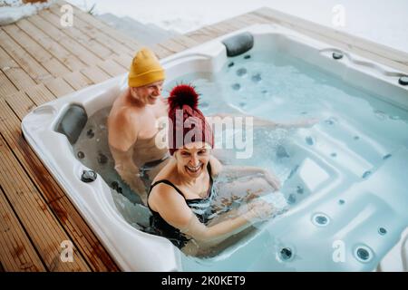 Das ältere Paar mit kitschiger Kappe genießt an kalten Wintertagen gemeinsam die Badewanne im Freien auf der Terrasse. Stockfoto