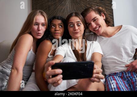 Glückliche junge multirassische Freunde in Hauskleidung, die Gesichter machen und Selfie auf dem Handy zu Hause machen Stockfoto
