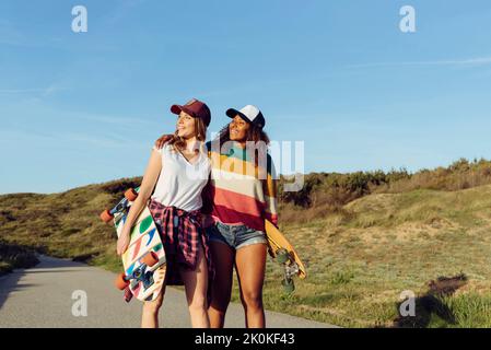 Zwei schöne, multiethnische, fröhliche Frauen, die Skateboards halten, während sie an einem sonnigen Tag mitten auf einer Straße spazieren gehen Stockfoto