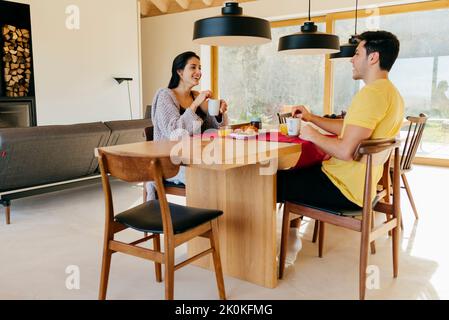 Attraktive hispanische glückliche Dame und junger Mann beim gemeinsamen Frühstück am Tisch in der modernen Küche Stockfoto