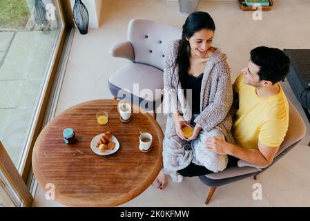 Attraktive hispanische glückliche Dame auf den Beinen des Mannes auf dem Stuhl im Zimmer und beim Frühstück Stockfoto