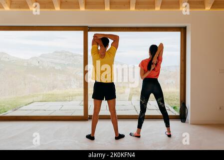 Rückansicht einer attraktiven Frau, die einen jungen Mann trägt, der die Arme an einem großen Fenster im Zimmer ausstreckt Stockfoto
