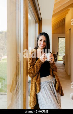 Attraktive hispanische Dame mit einem Becher Getränk, das im Zimmer steht und die Kamera anschaut Stockfoto