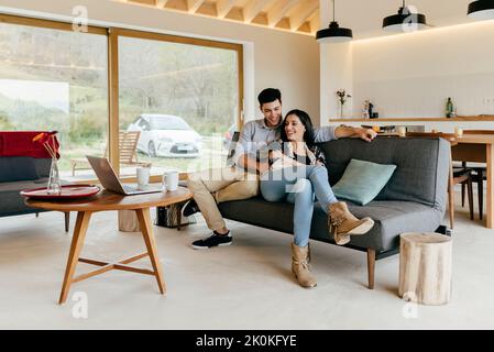 Junges attraktives Paar auf Sofa und Blick auf Laptop auf dem Tisch im Zimmer neben der Küche Stockfoto