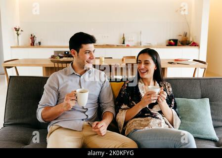 Fröhliches, attraktives Paar mit Becher, das auf einem Sofa in der Nähe der Küche sitzt Stockfoto