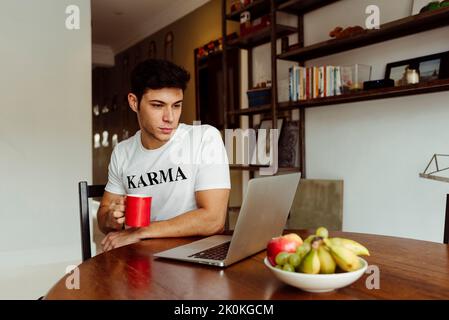 Gutaussehender kaukasischer Mann, der zu Hause Kaffee trinkt, während er an seinem Laptop auf dem Tisch arbeitet Stockfoto