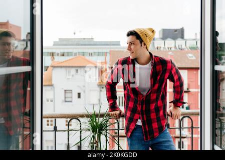 Trendiger, hübscher kaukasischer Mann mit Beanie-Hut und Hemd auf dem Balkon, der zu Hause wegschaut Stockfoto