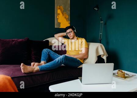 Attraktiver kaukasischer Mann, der mit geschlossenen Augen auf der Couch sitzt, während er zu Hause ein Nickerchen mit Musik über Kopfhörer macht Stockfoto