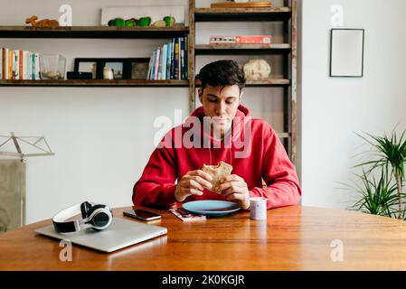 Ein gutaussehender, nachdenklicher kaukasischer Mann, der zu Hause isst, während er eine Mittagspause von der Arbeit mit einem Laptop auf dem Tisch hat Stockfoto