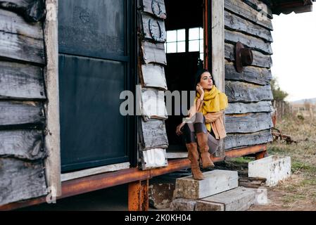 Stilvolle Frau in Stiefeln und Schal sitzen auf der Veranda des alten Holzhauses in der Landschaft mit Blick weg Stockfoto