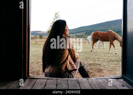 Rückansicht der stilvollen Frau in Stiefeln und Schal sitzen auf der Veranda des alten Holzhauses in der Landschaft weg schauen Stockfoto