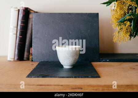 Weißer Keramiktopf auf schwarzem Schieferbrett in der Nähe alter Bücher und Strauß Mimosenblumen auf Holzregal Stockfoto