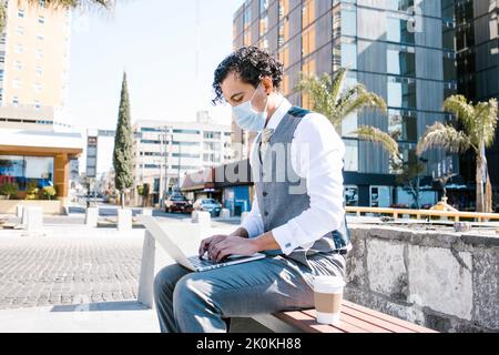 Selbstbewusster männlicher Unternehmer in elegantem Anzug und medizinischer Maske sitzt auf dem Bürgersteig mit Netbook und Kaffee zum Mitnehmen auf der sonnigen Straße während des Coronavirus p Stockfoto