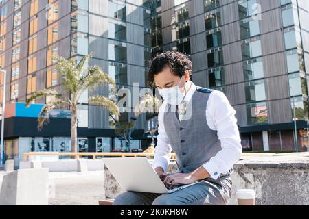 Selbstbewusster männlicher Unternehmer in elegantem Anzug und medizinischer Maske sitzt auf dem Bürgersteig mit Netbook und Kaffee zum Mitnehmen auf der sonnigen Straße während des Coronavirus p Stockfoto
