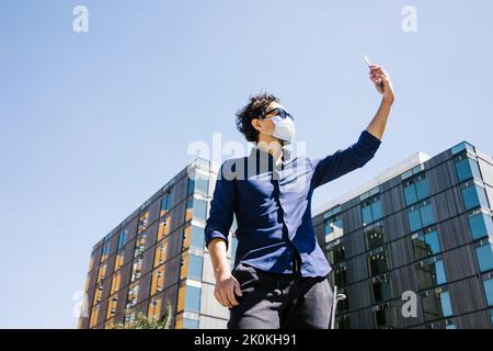 Von unten von männlich in elegantem Outfit und Schutzmaske, die Selbstporträt auf dem Smartphone, während auf der sonnigen Straße in der Nähe von modernen Gebäuden durin Stockfoto