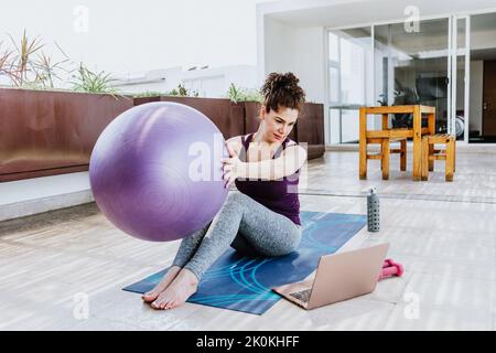 Ganzkörper einer jungen, sportlichen Frau, die auf einer Matte sitzt und den Ball in den Händen hält, während sie sich während des Ferntrainings auf Terra ein Tutorial-Video auf dem Laptop ansieht Stockfoto