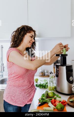 Seitenansicht einer positiven Frau, die frischen Sellerie in den Entsafter gab, während sie tagsüber in der Küche zu Hause gesunden Smoothie zubereitete Stockfoto