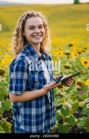 Lächelnd glücklich lockig Frau mit modernen Technologien für die Produktion von landwirtschaftlichen Produkten im Sonnenblumenfeld. Moderne Technologien in der Landwirtschaft Stockfoto