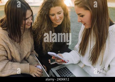 Eine Gruppe glücklicher Freundinnen in lässigen Outfits, die auf einer Steinbank auf dem Campus sitzen und gemeinsam auf einem Laptop und einem Smartphone surfen Stockfoto