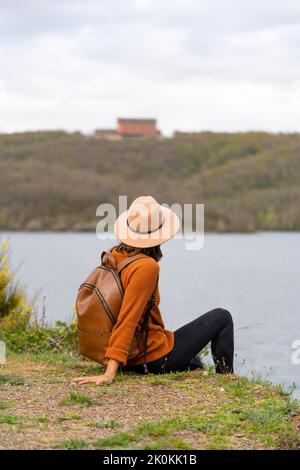 Seitenansicht einer nicht erkennbaren Frau mit Hut und Lederrucksack, die am Flussufer sitzt und wegschaut, während sie den Sommer in Palencia verbringt Stockfoto