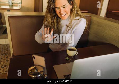 Von oben auf der jungen lächelnden Freiberuflerin, die im Café mit Tee und winkender Hand sitzt und einen Videoanruf führt, während sie am Laptop arbeitet Stockfoto