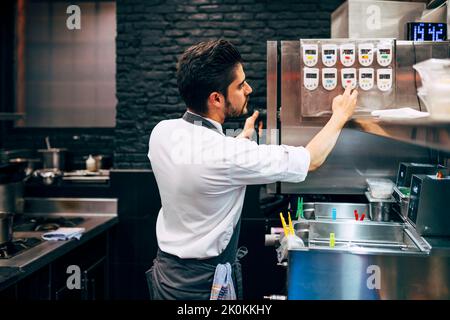 Seitenansicht des Mannes, der während der Arbeit in der Restaurantküche den Timer auf ein Metallgerät setzt Stockfoto