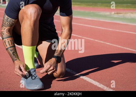 Seitenansicht eines anonymen männlichen Läufers, der auf der Rennstrecke sitzt und Schnürsenkel auf Turnschuhen bindet, während er sich im Sommer auf das Training im Stadion vorbereitet Stockfoto