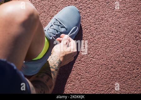 Draufsicht des anonymen männlichen Läufers, der auf der Rennstrecke sitzt und Schuhe an Turnschuhen bindet, während er sich im Sommer auf das Training im Stadion vorbereitet Stockfoto