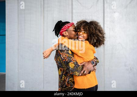 Seitenansicht liebevoller afroamerikanischer moderner Mann und Frau umarmen und lächeln hell an der grauen Wand auf der Straße Stockfoto